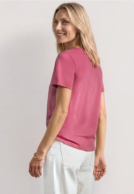 Eine lächelnde blonde Frau trägt ein pinkes T-Shirt und helle Jeans, sie blickt über die Schulter.