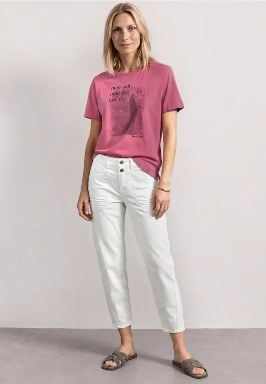 Frau in locker sitzendem pinkem T-Shirt mit Print, weißen Hosen und Sandalen vor hellem Hintergrund.