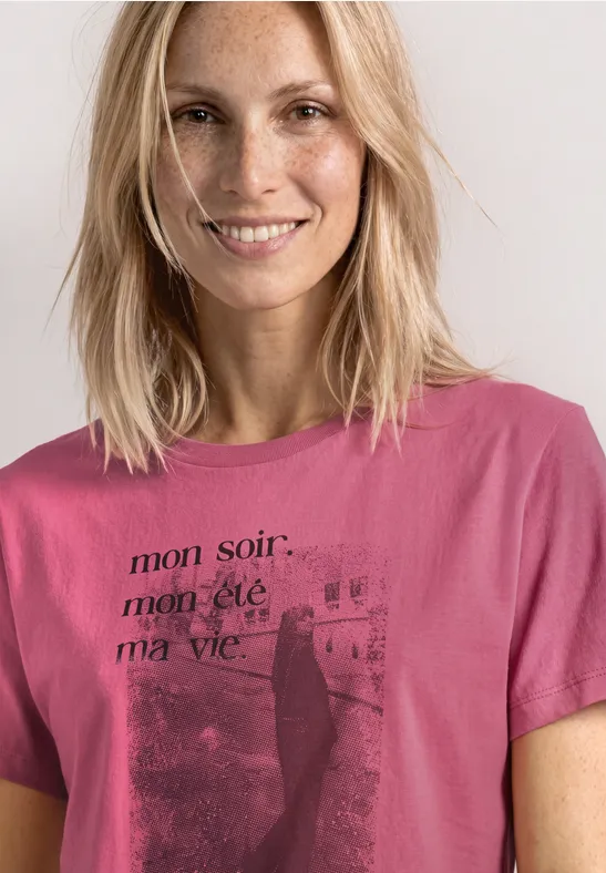 Lächelnde blonde Frau im pinken Shirt mit französischem Schriftzug vor hellem Hintergrund.