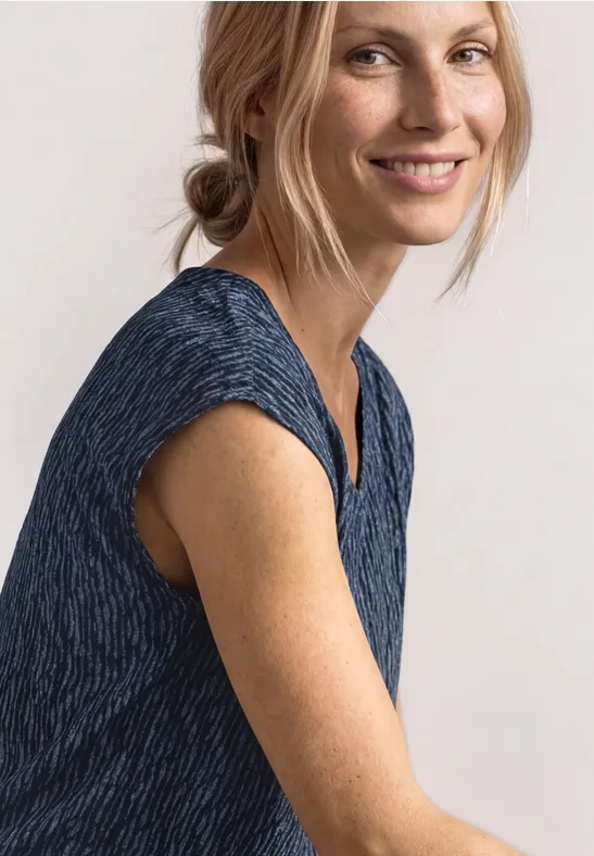 Vrouw in blauw mouwloos topje zittend opzij voor een lichte achtergrond.