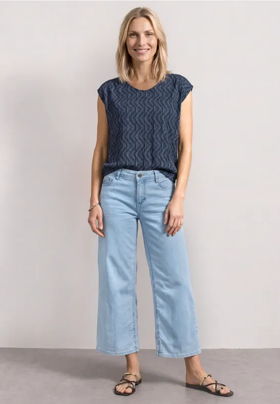 Vrouw voor lichte achtergrond, met blauw gebreid topje, wijde lichtblauwe jeans en zwarte sandalen.