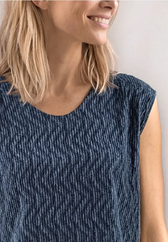 Een vrouw met halflang blond haar draagt een blauw patroon-T-shirt en glimlacht zijwaarts voor een witte achtergrond.