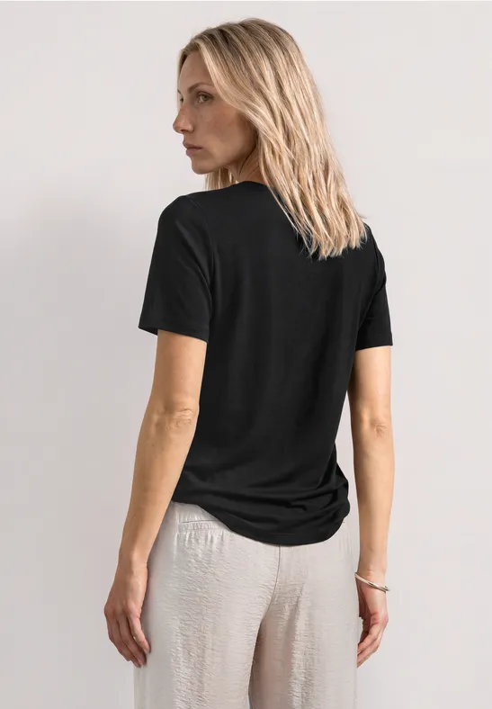 Eine Frau steht von hinten betrachtet, trägt ein schwarzes T-Shirt und helle Hose.