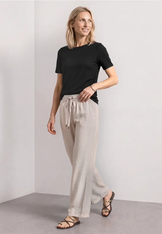 Frau in schlichtem Raum trägt schwarzes T-Shirt, helle Leinenhose mit Kordelzug und schwarze Sandalen.