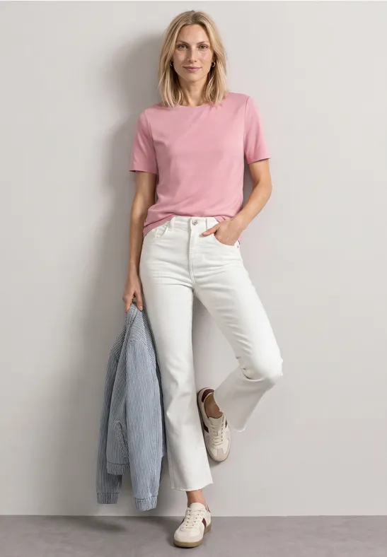 Frau in rosa T-Shirt und weißen Jeans lehnt lässig an der Wand, hält eine Jeansjacke in der Hand.
