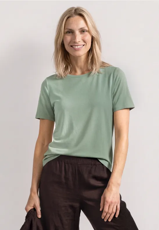 Vrouw draagt een simpel mintgroen T-shirt en een bruine linnen broek tegen een lichte, rustige achtergrond.