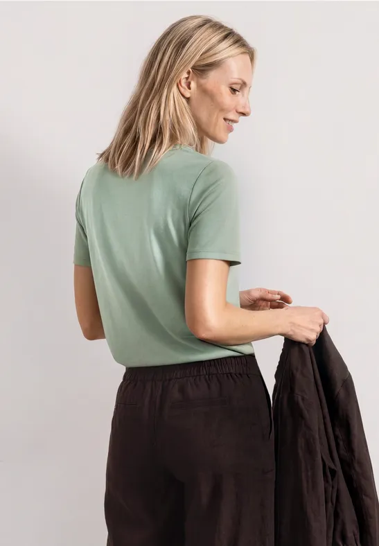 Blonde vrouw van achteren in mintgroen t-shirt, die een zwarte jas vasthoudt.