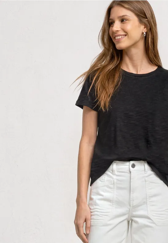 Eine Frau in schwarzem T-Shirt und weißer Hose vor hellem Hintergrund.