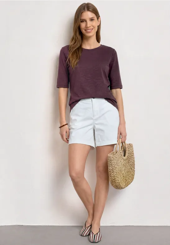 Frau in lila Shirt, weißen Shorts und Strohtasche vor weißem Hintergrund.