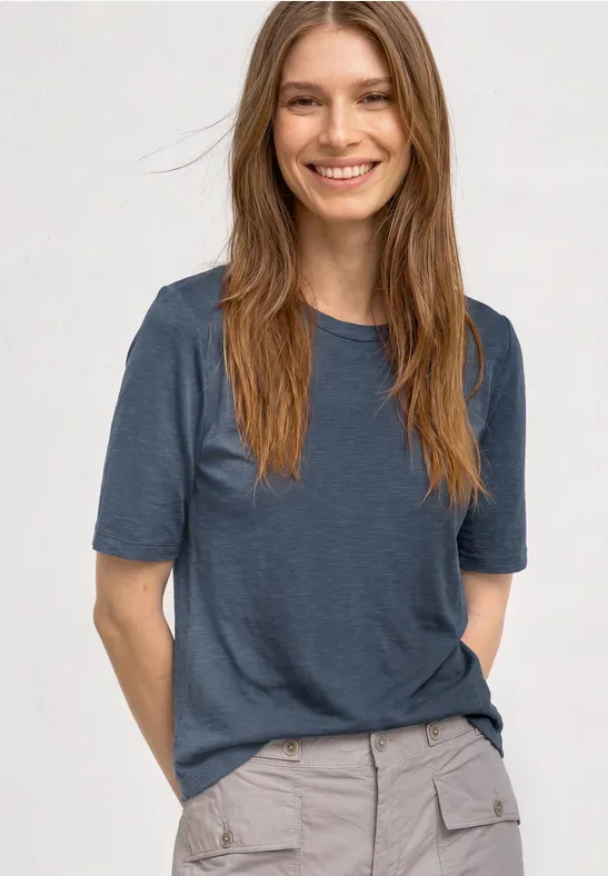 Frau mit blauem T-Shirt und grauer Hose lächelt vor weißem Hintergrund.