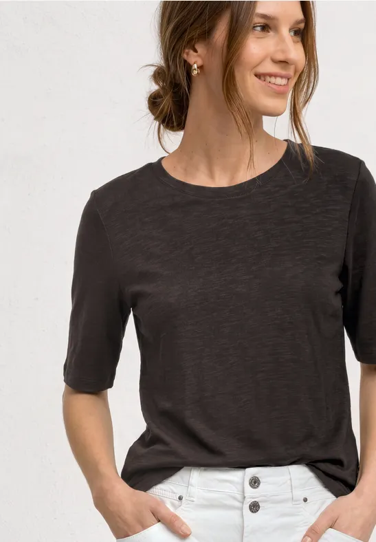 Frau trägt dunkelgraues T-Shirt und weiße Hose mit Knöpfen, Hände in den Taschen.