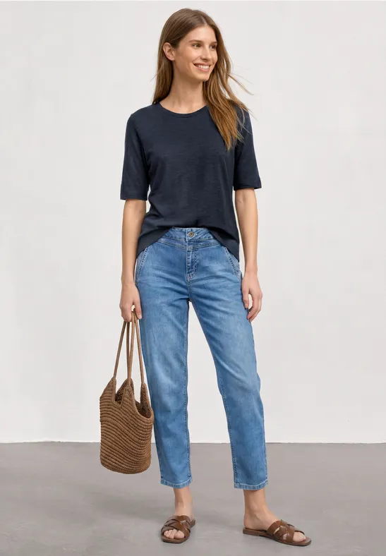 Frau in blauem T-Shirt, weiten Jeans und Sandalen, trägt eine braune Korbtasche vor neutralem Hintergrund.