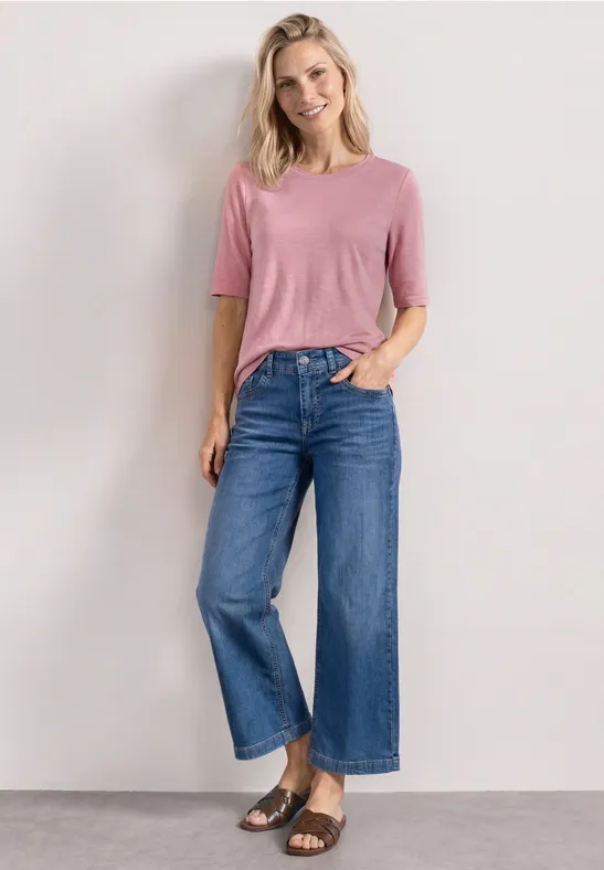 Model draagt een roze t-shirt, blauwe wijde high-waist jeans en zwarte sandalen voor een neutrale achtergrond.