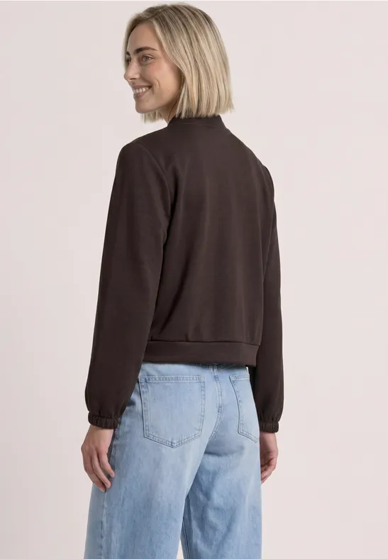 Vrouw lacht in een bruin truitje met jeans, kijkt over haar schouder.