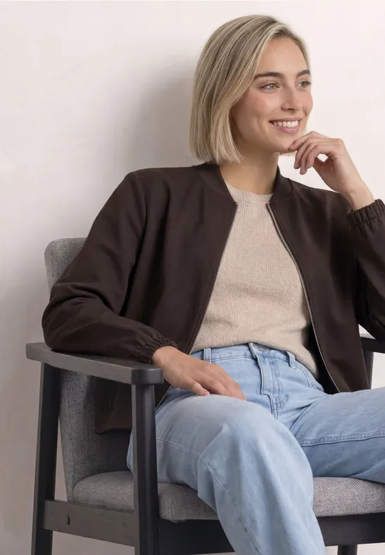 Een jonge blondine zit glimlachend op een grijze fauteuil, draagt een beige top met bruine cardigan en blauwe spijkerbroek.