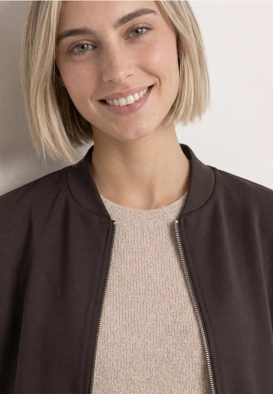 Een vrouw met een kort blond bobkapsel, draagt een donkere jas over een licht truitje en lacht.