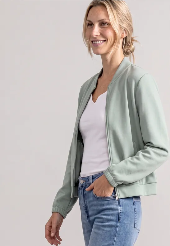Frau trägt hellgrüne Bomberjacke, weißes Top und Jeans, lächelt entspannt