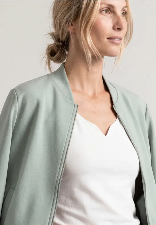 Blonde Frau in weißem Shirt und mintgrünem Blouson, seitlich in die Ferne blickend.