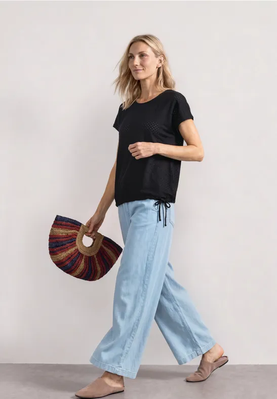 Frau in schwarzem T-Shirt, hellblauer weiter Hose und Sandalen, die eine fächerförmige Tasche hält.