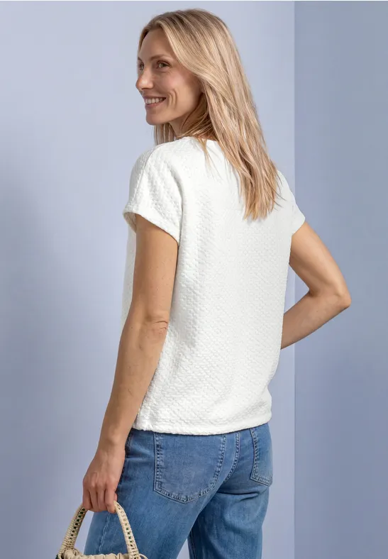 Eine blonde Frau steht seitlich, trägt ein weißes Shirt und Jeans, hält eine Strohtasche und lächelt.