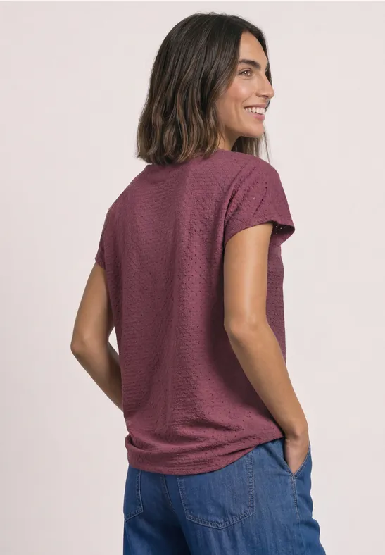 Eine Frau von hinten in einem kurzärmligen, strukturierten T-Shirt in Dunkelrot und blauen Jeans.