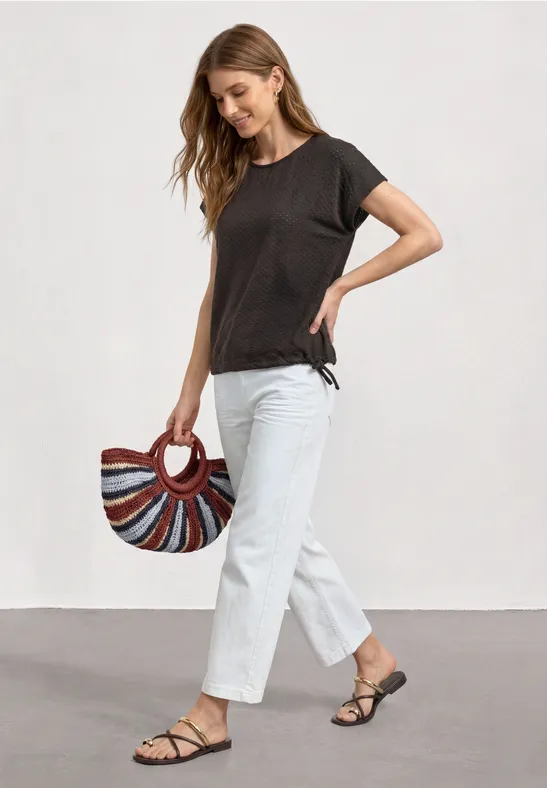 Eine Frau trägt weiße Hosen, schwarzes T-Shirt und Sandalen und hält eine bunte, rund gestreifte Handtasche.