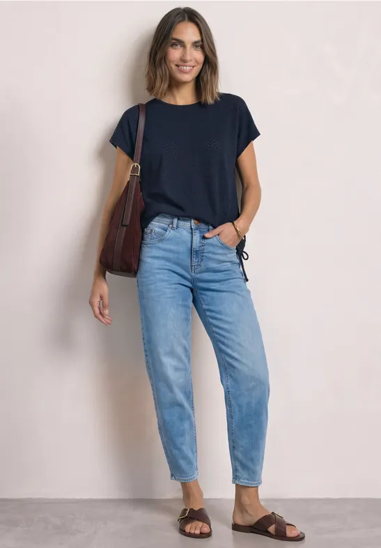 Frau vor hellem Hintergrund in dunkelblauem T-Shirt, Jeans, braunen Sandalen und Schultertasche, sie lächelt.