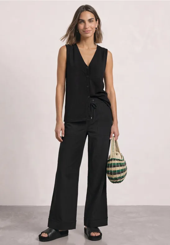 Femme en top sans manches noir et pantalon large, tient un sac en osier, porte des sandales.