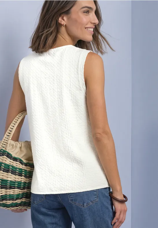 Frau in weißem, ärmellosem Top und Jeans, trägt handgewebte Tasche, blickt seitlich und lächelt.