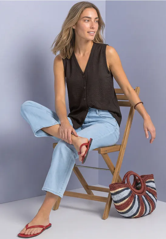 Frau in schwarzem Top, hellen Jeans und roten Sandalen sitzt lässig auf einem Holzstuhl, Korbtasche daneben.