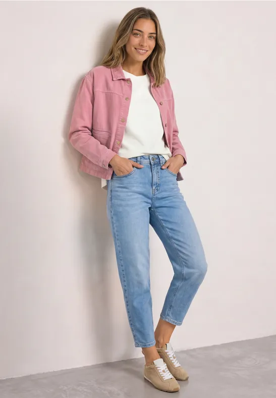 Eine Frau trägt rosa Jacke, weißes Shirt, blaue Jeans und beigen Sneaker vor hellem Hintergrund.