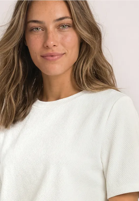 Porträt einer lächelnden Frau mit braunen Haaren in einem schlichten weißen Kurzarmshirt vor hellem Hintergrund.