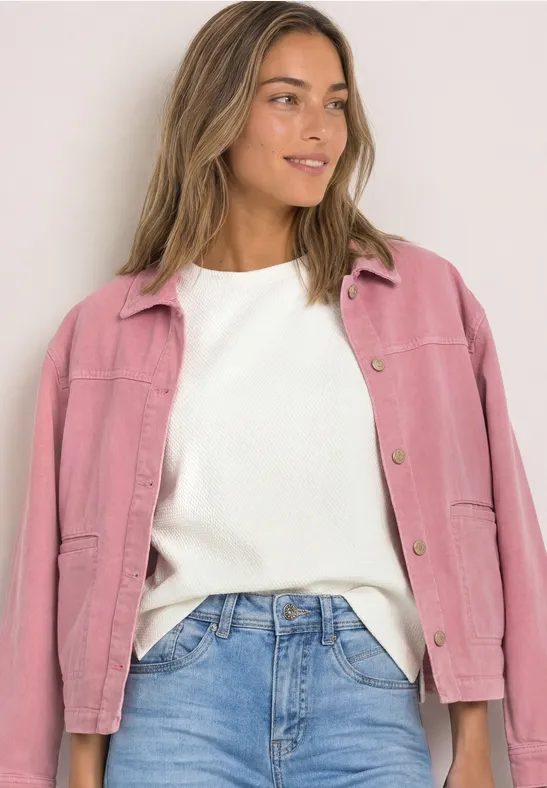 Frau in blauer Jeans, weißem Pullover und pinker Jacke, die lächelnd seitlich posiert.