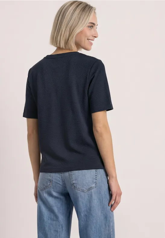Frau mit kurzem blondem Haar trägt schwarzes T-Shirt und blaue Jeans, Rückenansicht.