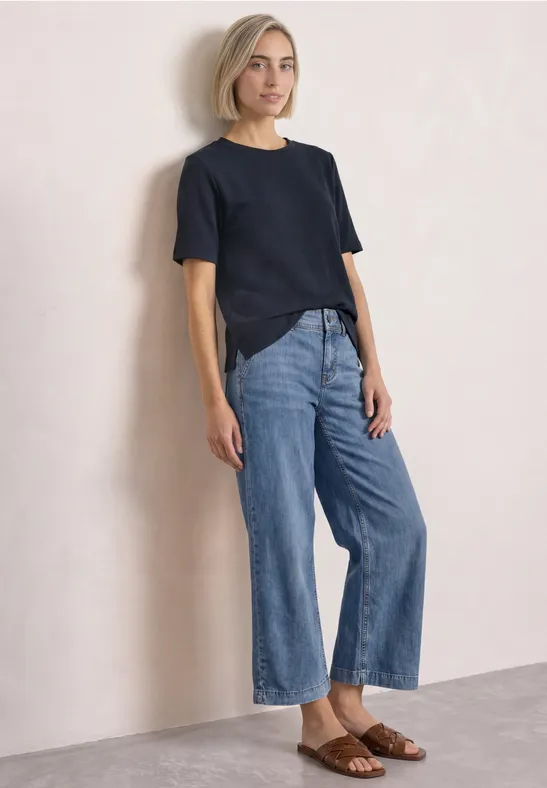 Frau in schwarzem T-Shirt, weiten Jeans und braunen Sandalen vor hellem Hintergrund.