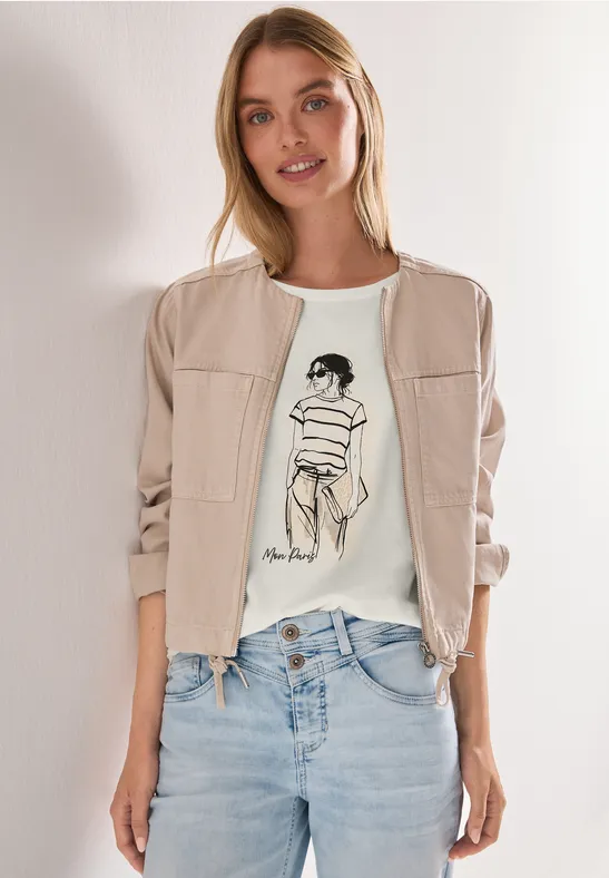 Vrouw in blauwe jeans, wit t-shirt met schetsprint en beige bomberjack voor witte achtergrond.
