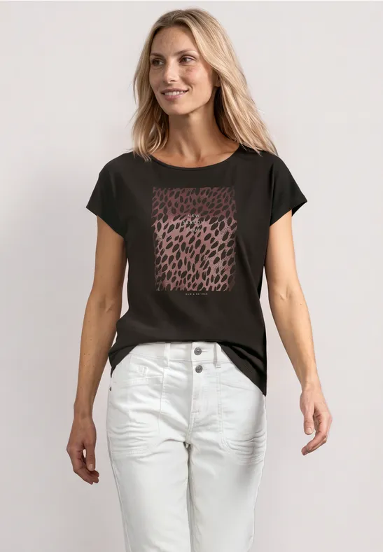 Frau in schwarzem T-Shirt mit Leopardenprint und weißen High-Waist-Hosen vor hellem Hintergrund