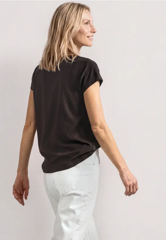 Frau von hinten in schwarzem T-Shirt und weißer Hose vor hellem Hintergrund.