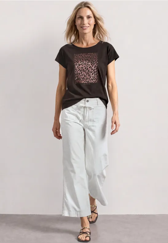 Frau in schwarzem T-Shirt mit Leoparden-Print, weiten weißen 7/8-Jeans und schlichten Sandalen steht lässig da.