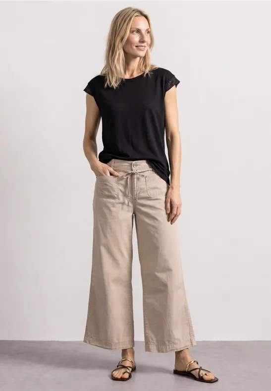 Een slanke vrouw in een zwart T-shirt, beige wijdvallende broek en sandalen, ontspannen met handen in de zakken.