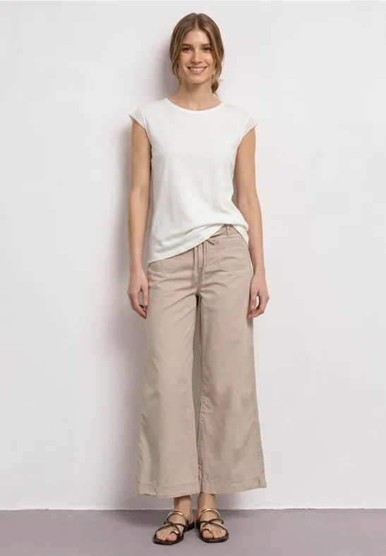 Junge Frau in weißem T-Shirt, beigen Culotte-Hosen und Sandalen vor neutralem Hintergrund stehend.