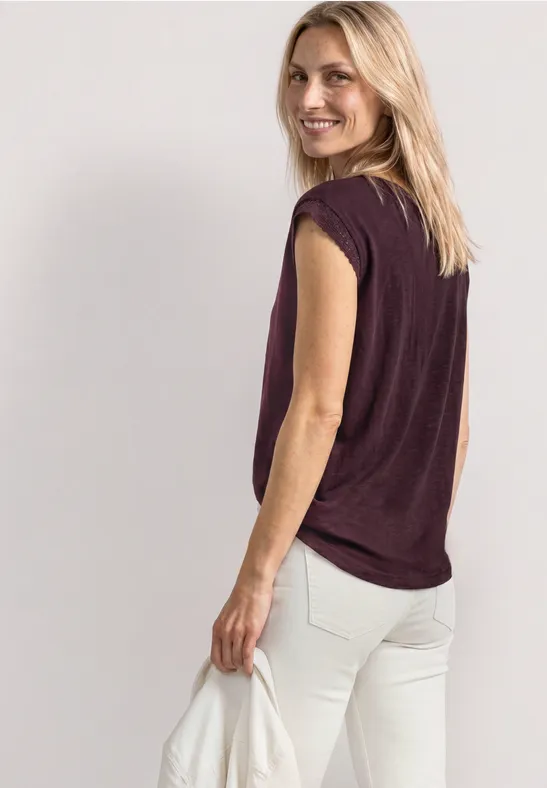 Vrouw met blond haar in zijaanzicht, gekleed in een mouwloze paarse top en lichte broek.