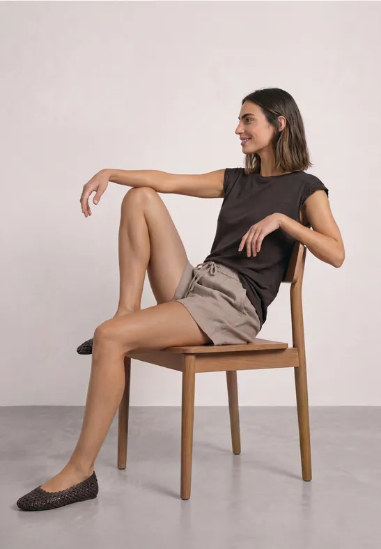Vrouw zit ontspannen opzij op een houten stoel, draagt een zwart shirt en beige short.