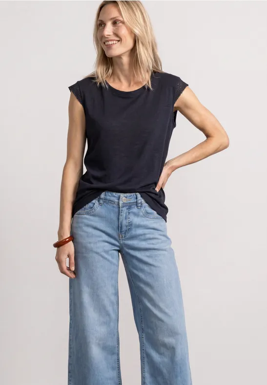 Vrouw in een zwart t-shirt en lichtblauwe, wijde jeans poseert voor een neutrale achtergrond.