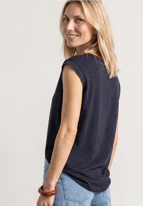 Een vrouw van achteren draagt een zwart mouwloos topje en blauwe jeans, blond haar en een houten armband.