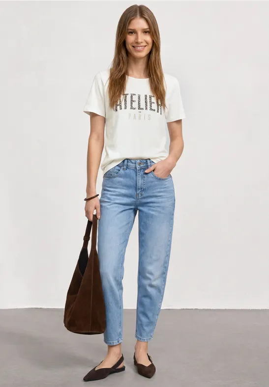 Frau in hellblauen Jeans und weißem 'Atelier'-T-Shirt, schwarze Ballerinas und braune Schultertasche.