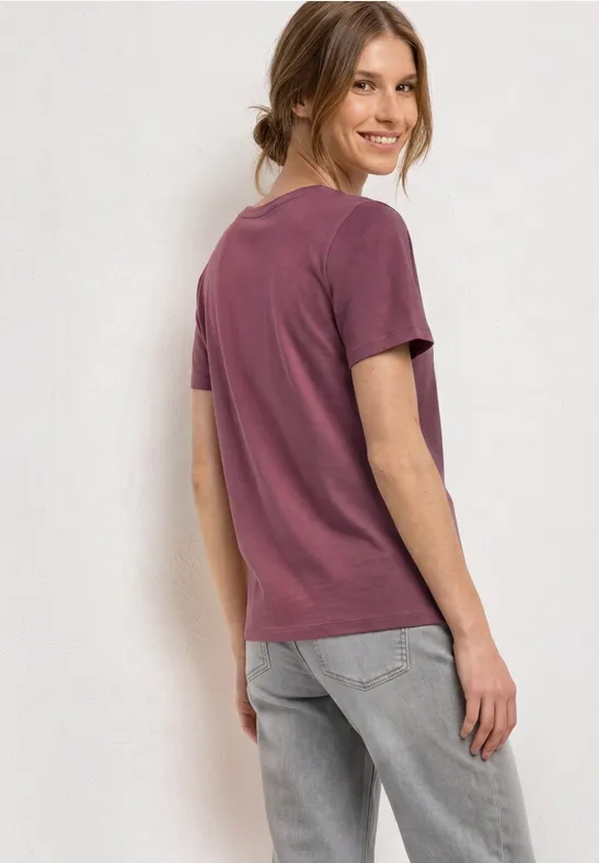 Vrouw in paars T-shirt en grijze spijkerbroek staat zijwaarts voor neutrale achtergrond