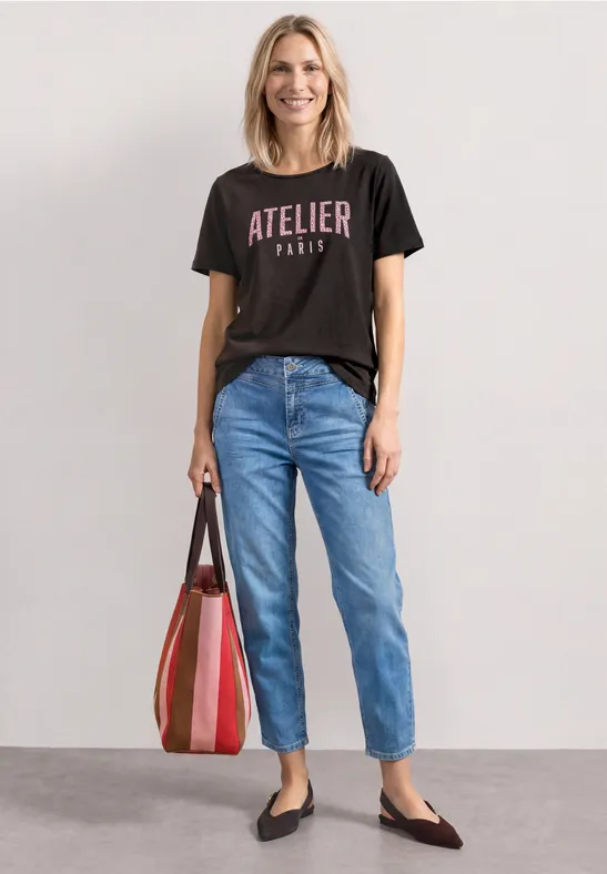 Femme porte un t-shirt noir «Atelier Paris», jean bleu, ballerines noires et grand cabas rayé.