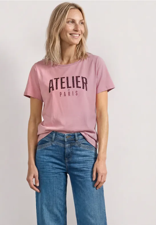 Persoon draagt roze T-shirt met opdruk 'ATELIER PARIS' en een blauwe jeans.