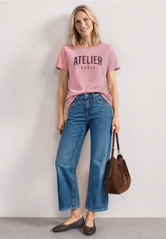 Vrouw in roze 'ATELIER PARIS' T-shirt, wijd jeans, mocassins en bruine tas tegen een witte muur.